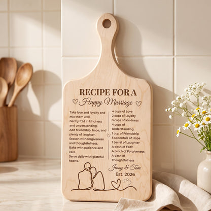 A personalized wooden recipe for a happy marriage cutting board with a handle, standing on a marble countertop in a kitchen setting. The board is engraved with a romantic poem, the names 'Jerry & Jim', and 'Est.