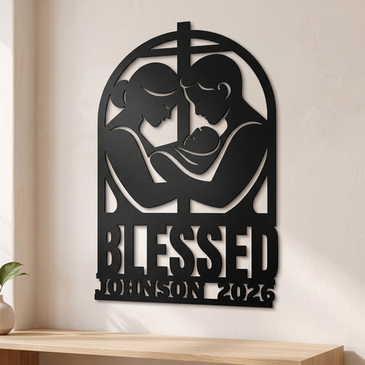 Black metal personalized wall art featuring a silhouette of a couple holding a baby under a cross, with the text 'BLESSED JOHNSON 2026' at the bottom, displayed on a beige wall above a wooden shelf.