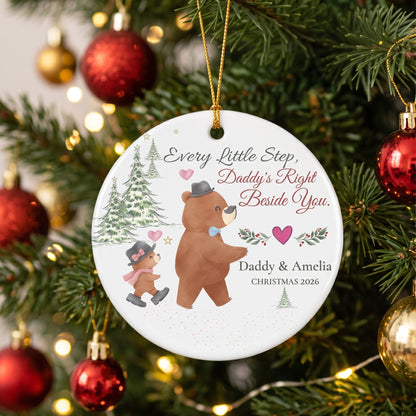 A personalized round Christmas ornament hanging on a decorated Christmas tree. The white ornament features an illustration of a teddy bear and a child, along with the text 'Every Little Step, Daddy's Right Beside You.